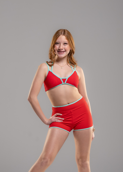 A young woman with red hair posing in a red two-piece swimwear set with light blue trim, consisting of a strappy bikini top and matching shorts, against a solid gray background.