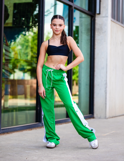 Woman in black sports bra and green track pants with white stripes standing outdoors.