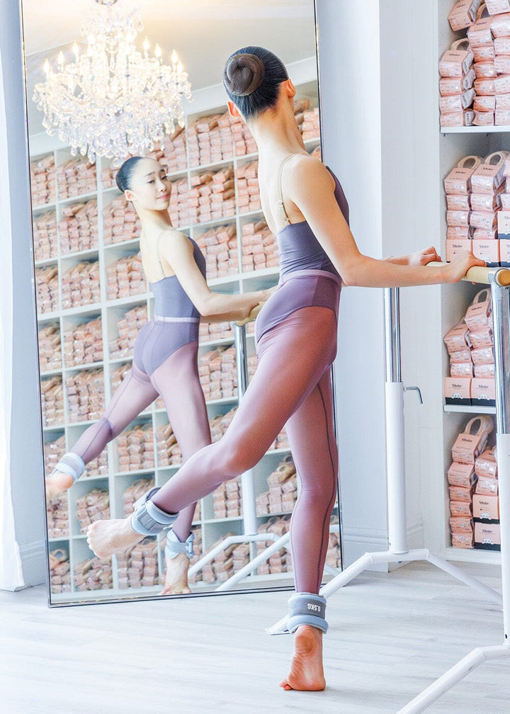 A ballet dancer in a purple leotard is performing a dance move with one leg lifted and an ankle weight on the other leg, standing on a barre with pink wraps in the background.