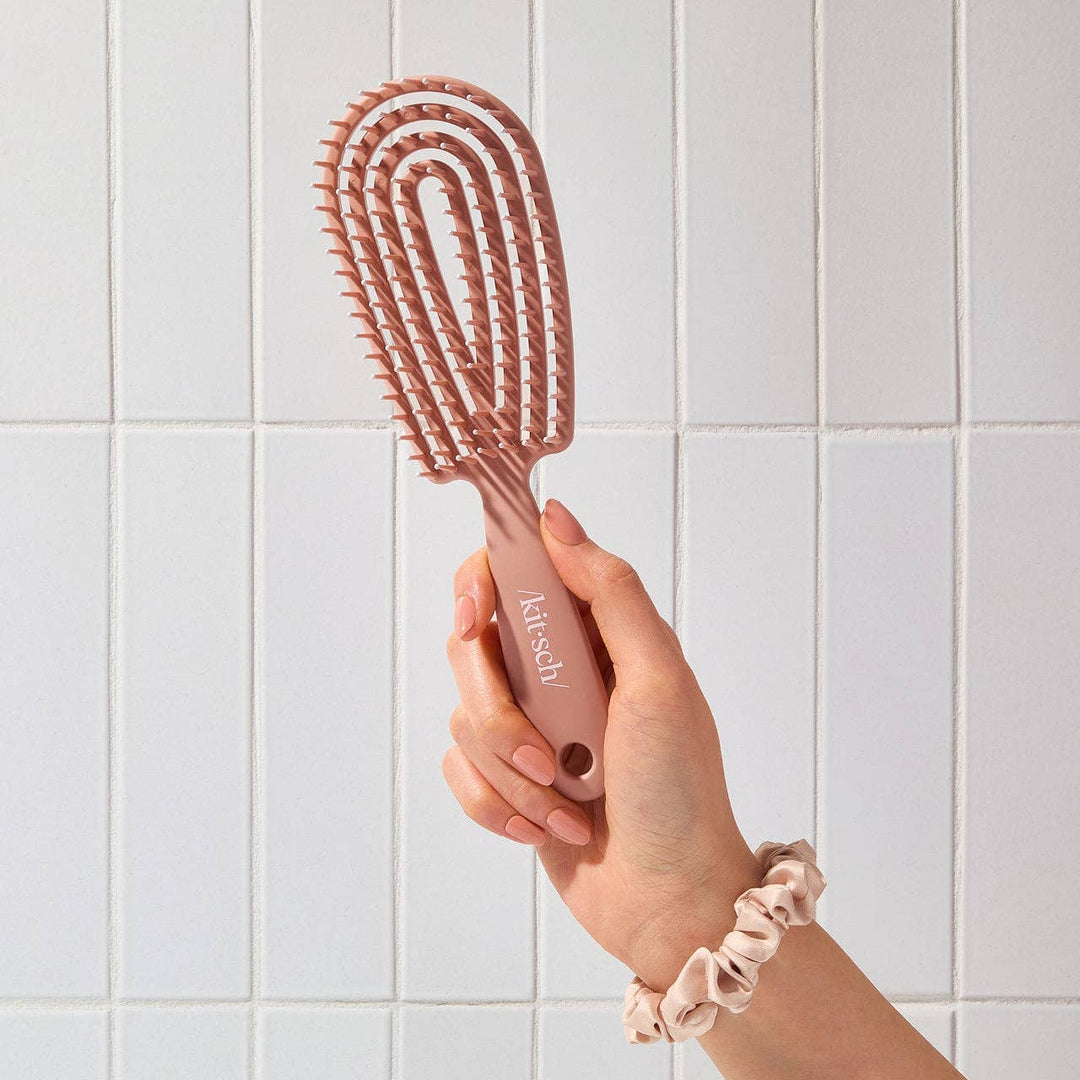 Hand holding a pink hairbrush with 'Kissch' branding against a tiled wall background