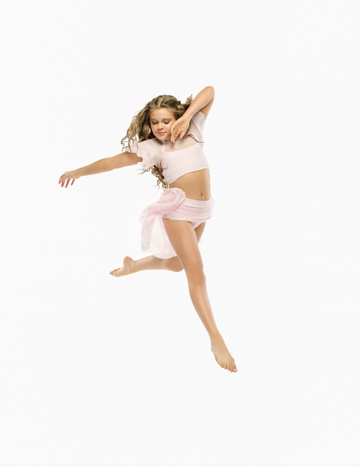 A young girl with curly brown hair is captured mid-air in a dance pose, wearing a light pink two-piece outfit consisting of a short-sleeve crop top and a matching skirt tied at the waist, set against a solid white background.