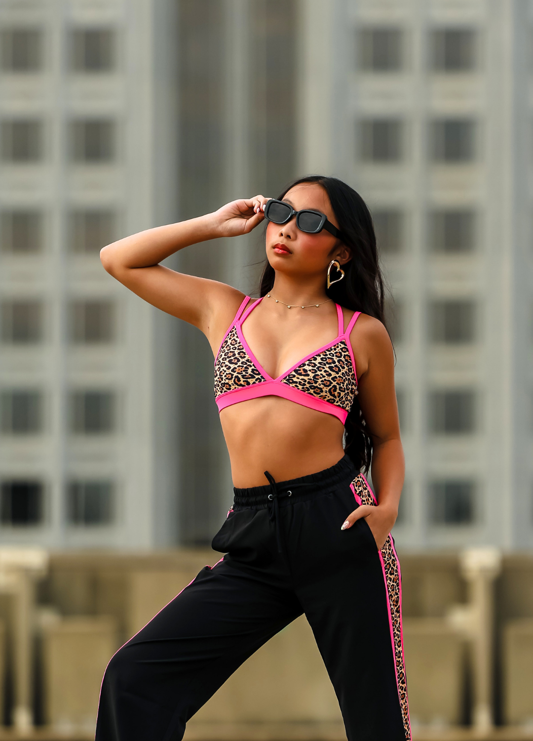 Woman wearing a leopard print bikini top and black pants with pink trim against a blurred cityscape background.