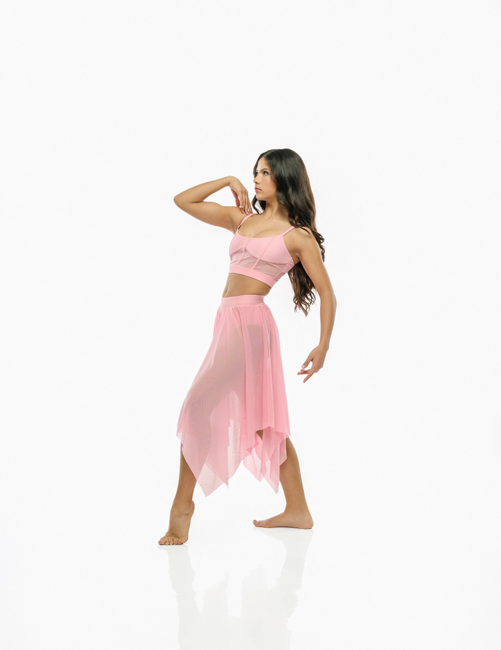 A woman modeling a light pink two-piece dance or yoga set consisting of a crop top and an asymmetrical skirt, posed against a seamless white background.