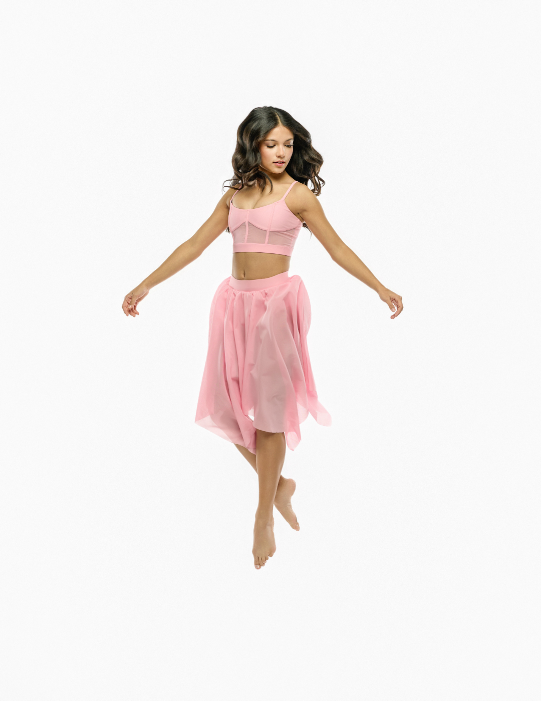 A woman floating mid-air in a light pink two-piece dance or yoga set, featuring a strappy crop top and a sheer flowy skirt, against a solid white background.