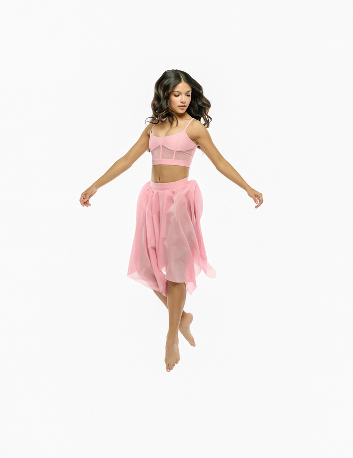 A woman floating mid-air in a light pink two-piece dance or yoga set, featuring a strappy crop top and a sheer flowy skirt, against a solid white background.