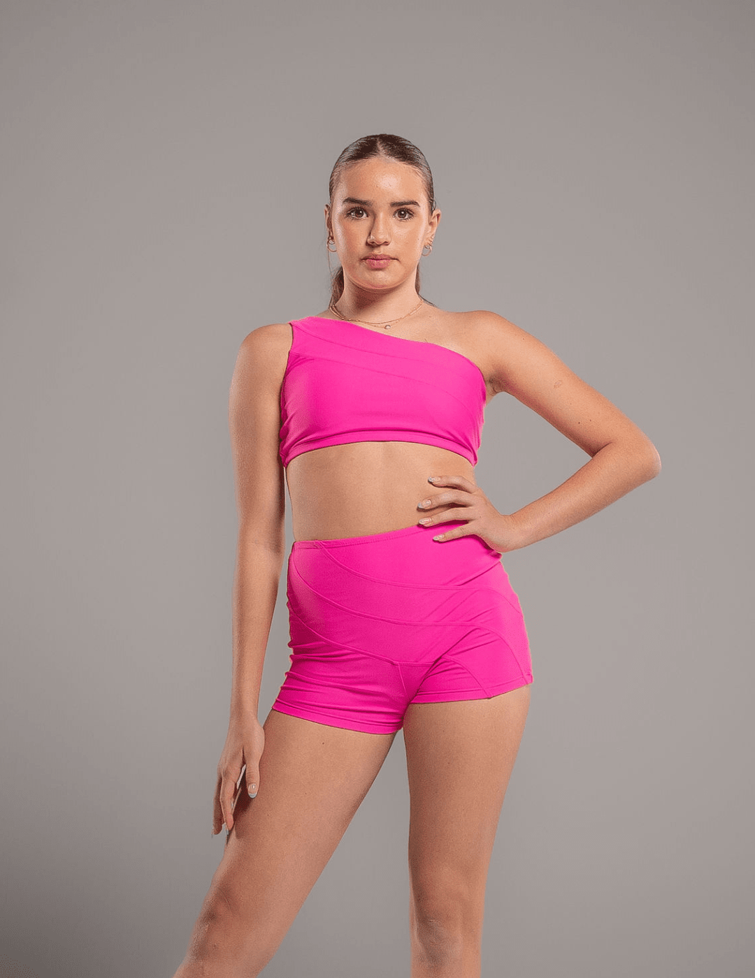 Woman wearing a bright pink one-shoulder top and shorts on a gray background