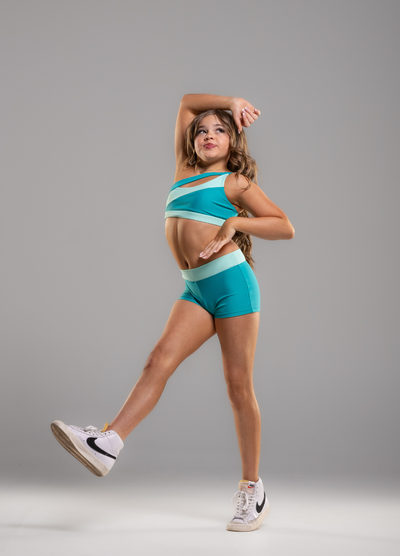 A woman posing in a matching turquoise and white athletic set consisting of a sports bra and shorts, paired with white Nike sneakers, against a solid gray studio background.
