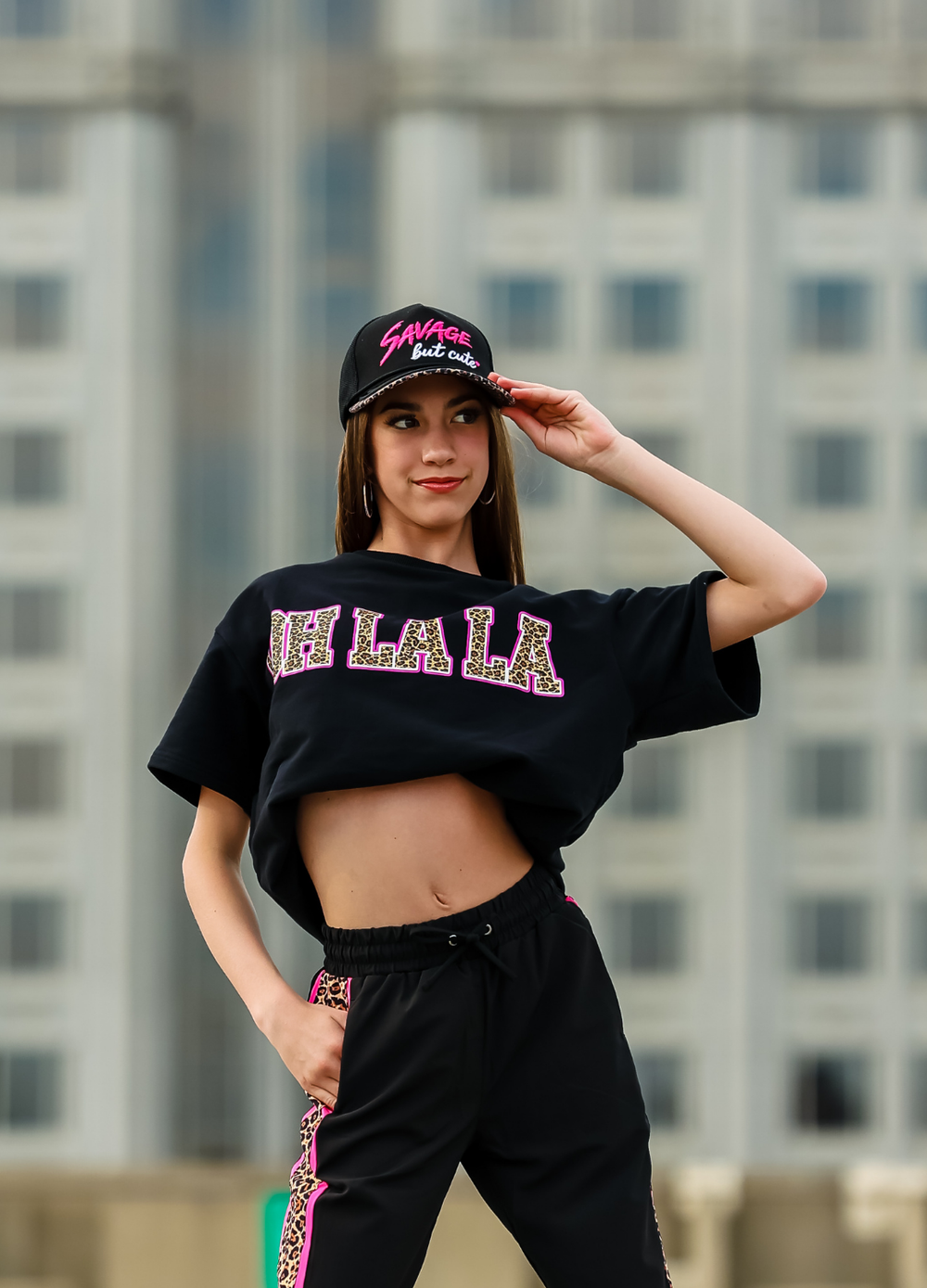 Person wearing a black crop top with 'OH LA LA' text and a cap, standing in front of a blurred building background.