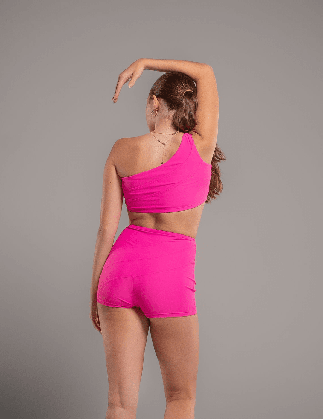 Person wearing a bright pink one-shoulder top and shorts set on a gray background
