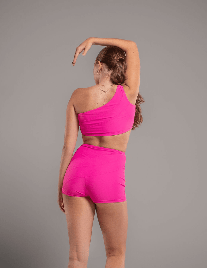Person wearing a bright pink one-shoulder top and shorts set on a gray background