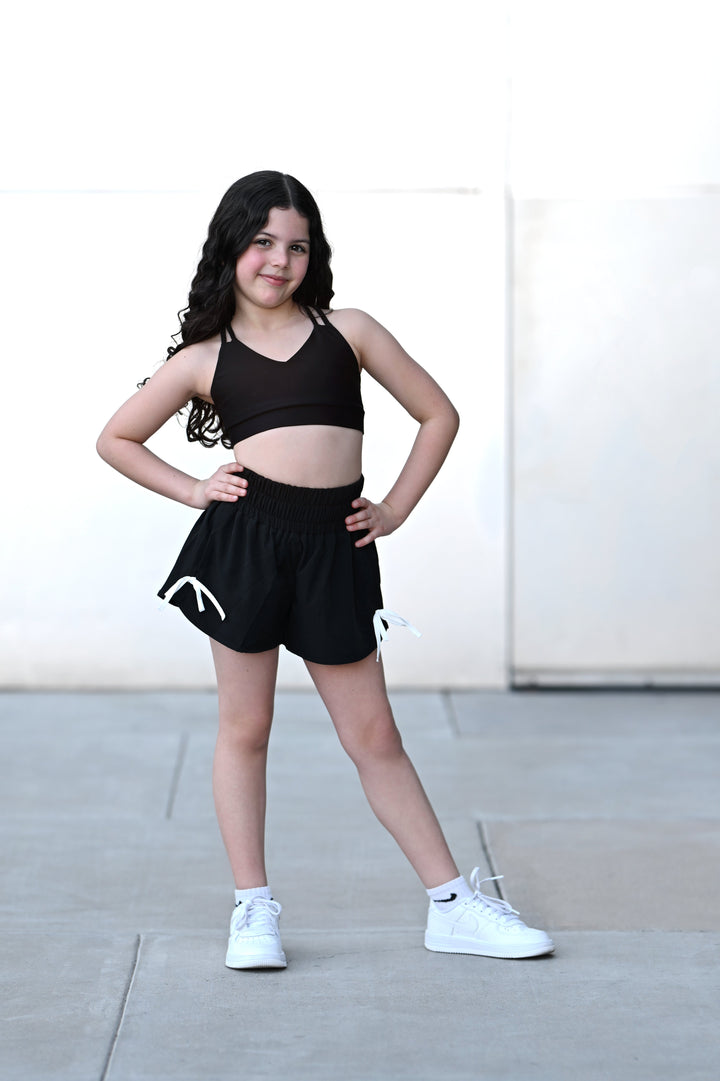 Young girl in black athletic outfit with white sneakers standing on a sidewalk.