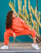 Woman in bright orange tracksuit squatting in front of a cactus plant