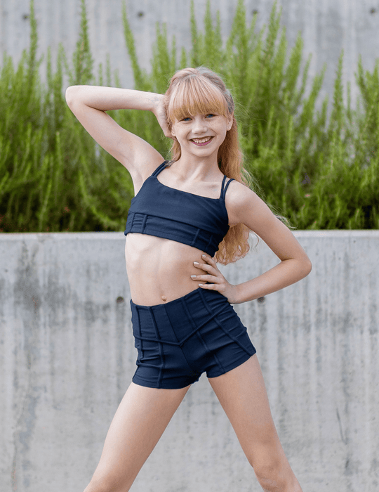 Young girl in navy sports bra and shorts posing outdoors with greenery in the background
