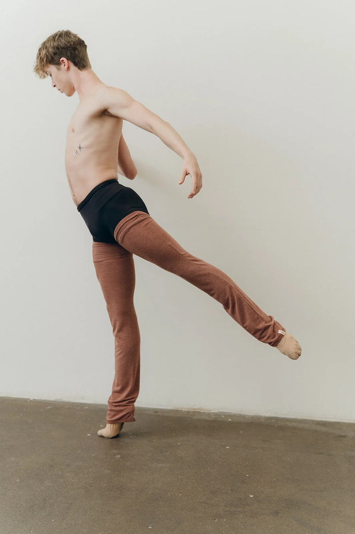 A shirtless male dancer in a studio, wearing black briefs and rust-brown leggings, balancing on one foot with one leg extended backward.
