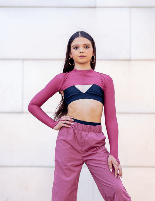 Woman wearing a pink long-sleeve top and pink pants with a black crop top underneath, standing against a light-colored wall.