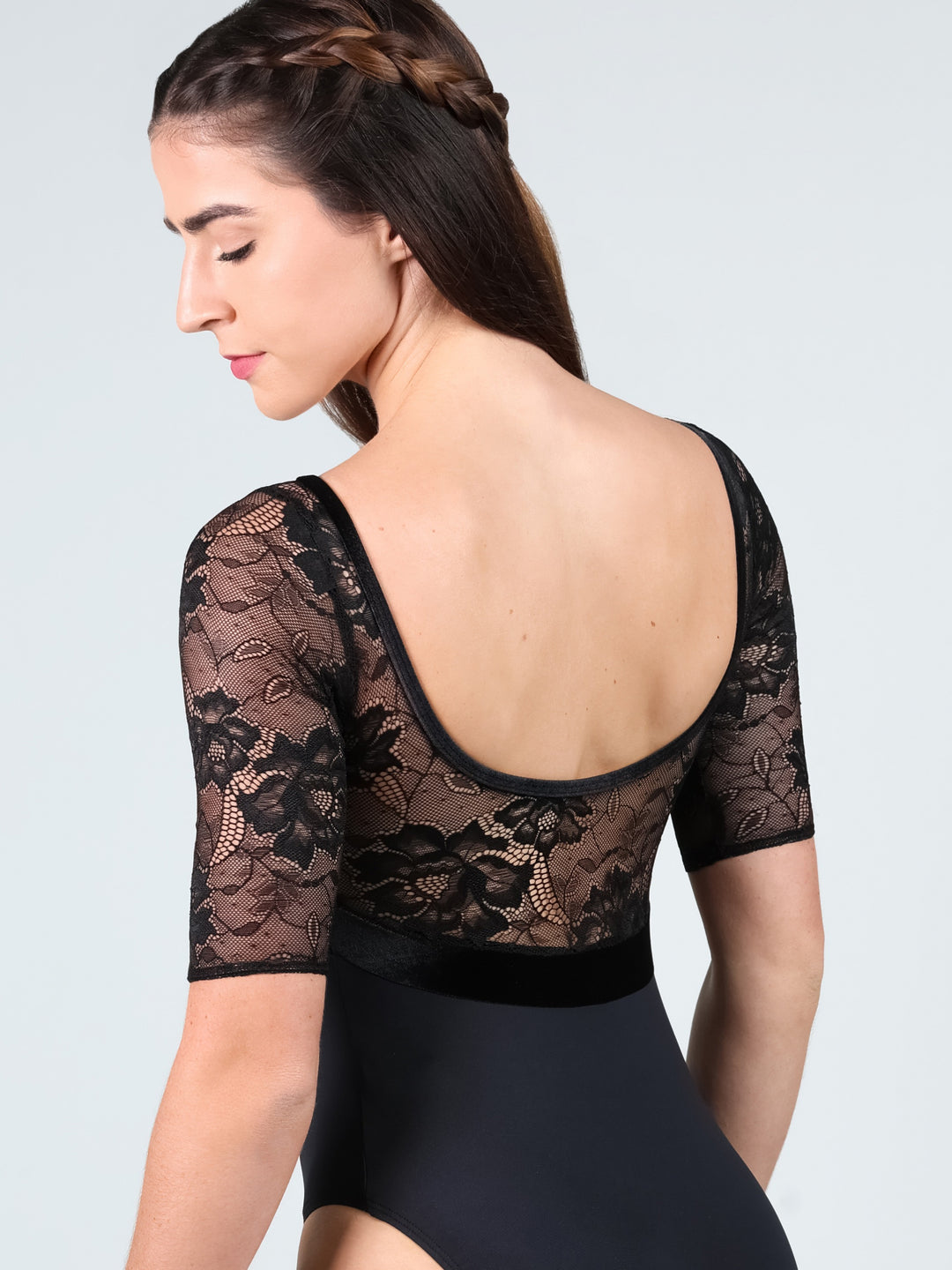 A woman shown from the back wearing a black lace leotard with short sleeves and a solid black bodysuit underneath, against a light gray studio background.