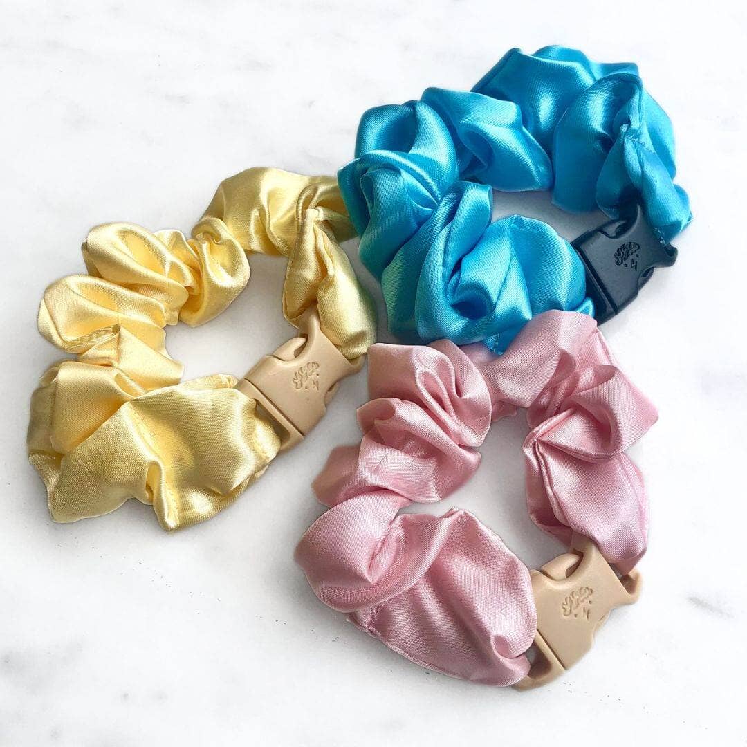 Three satin scrunchies in gold, blue, and pink on a white background