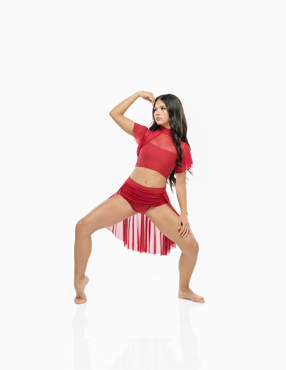 A woman modeling a red two-piece dance outfit consisting of a sheer mesh short-sleeve crop top with fringe trim and matching red shorts with a striped fringe hem, posed against a seamless white background.