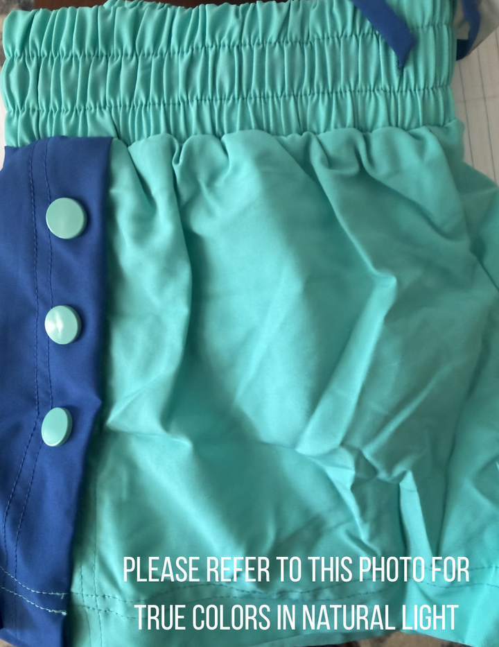 Teal shorts with buttons on a blue background, text overlay 'Please refer to this photo for true colors in natural light'.