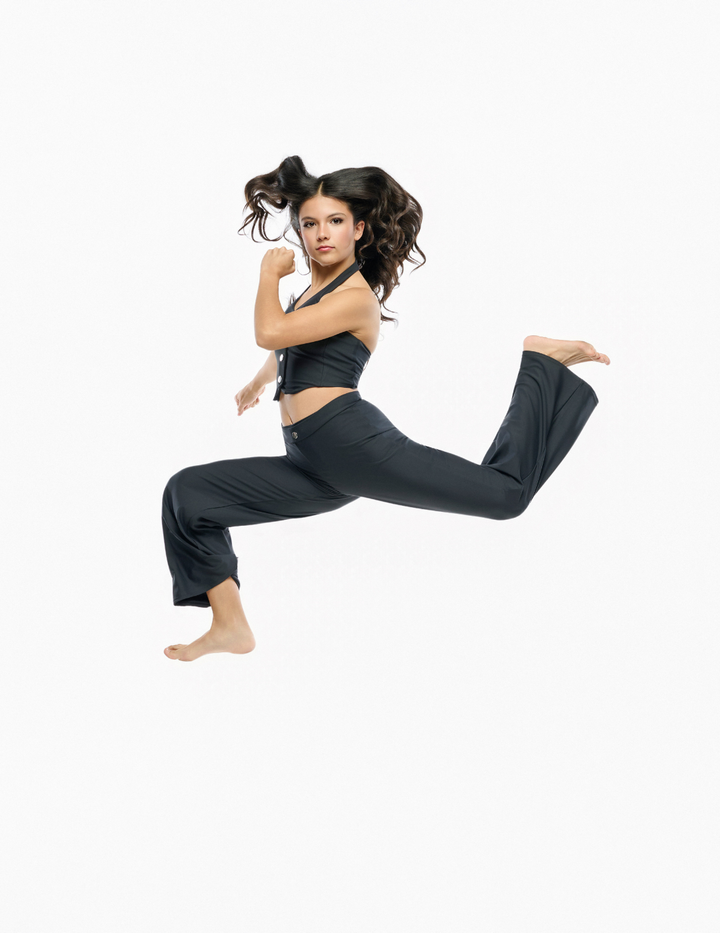 A woman in a black athletic crop top and matching wide-leg pants jumps in a dynamic lunge pose against a solid white background.
