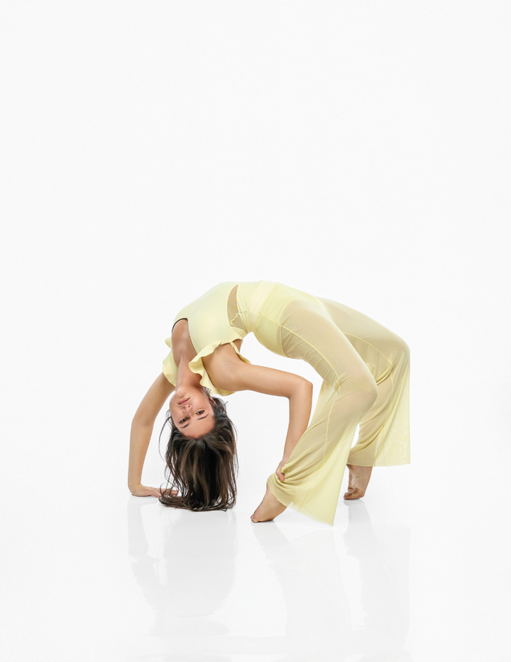 A woman in a pale yellow sleeveless top and matching wide-leg pants poses in a deep backbend on a seamless white studio background.