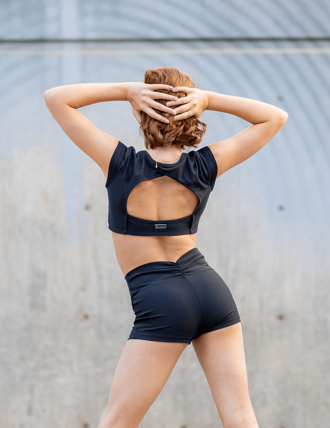 Person wearing a black sports bra and shorts with a visible brand logo, standing against a gray wall.