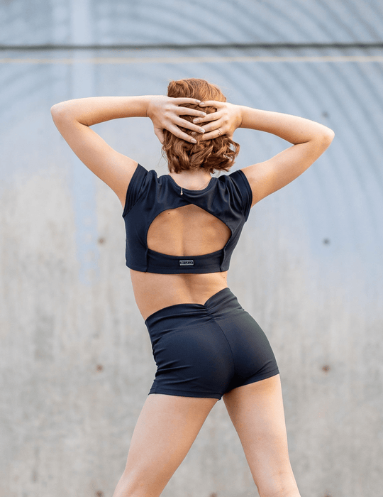 Person wearing a black sports bra and shorts with a visible brand logo, standing against a gray wall.