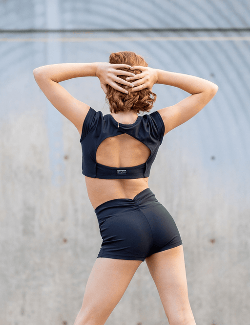 Person wearing a black sports bra and shorts with a visible brand logo, standing against a gray wall.