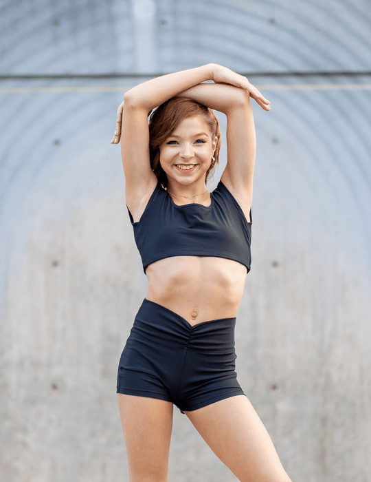Woman in black athletic wear stretching outdoors against a gray wall.