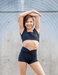 Woman in black athletic wear stretching outdoors against a gray wall.