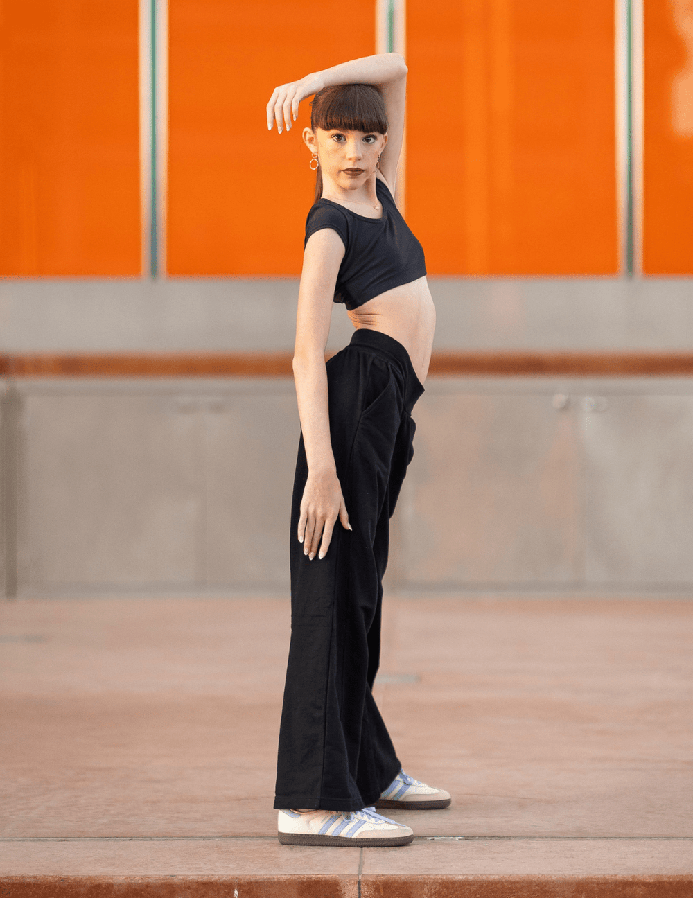 Person wearing a black outfit with high-waisted pants and sneakers against an orange wall.