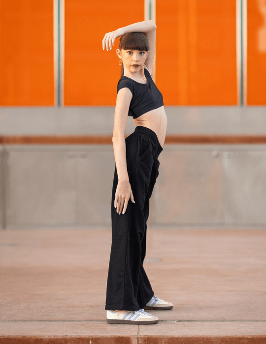 Person wearing a black outfit with high-waisted pants and sneakers against an orange wall.
