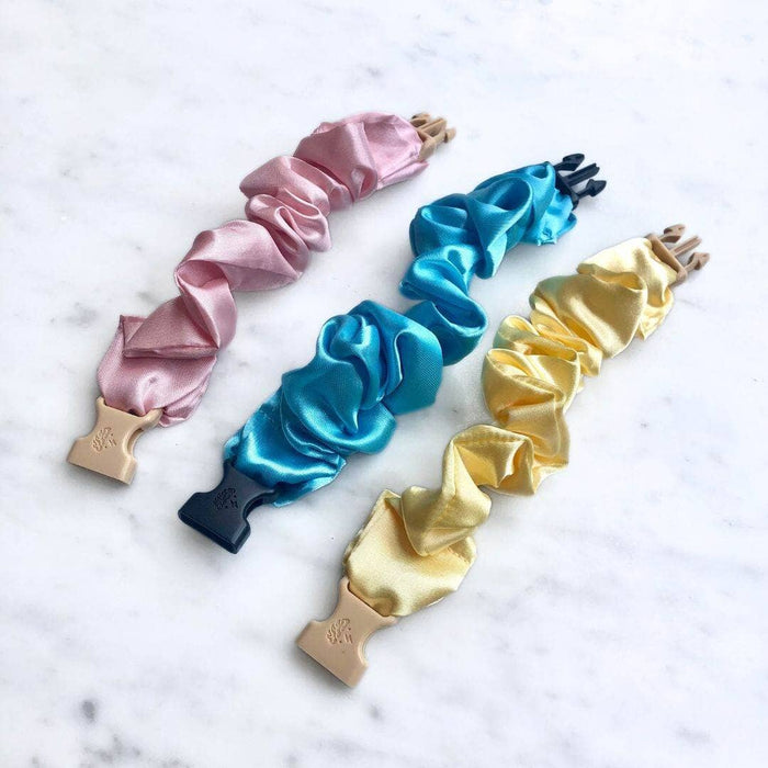 Set of four satin hair scrunchies in pink, blue, gold, and black on a marble surface.
