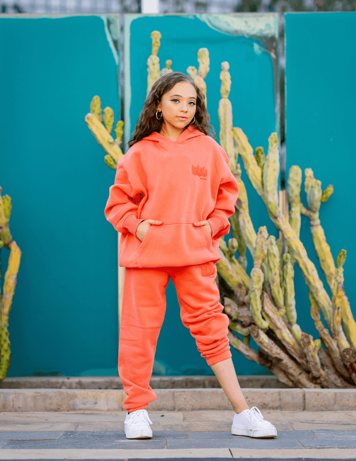 Person wearing a bright orange hoodie and pants standing in front of a teal wall with cacti.