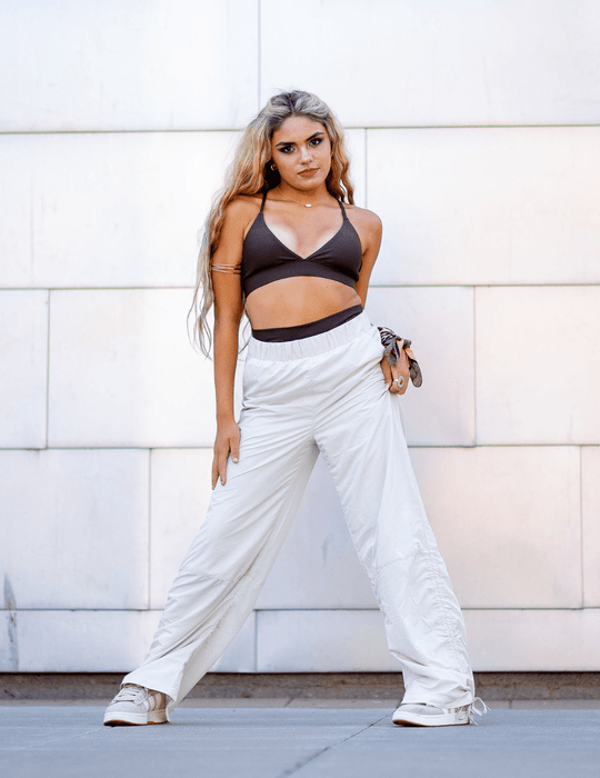 Woman wearing a black crop top and white wide-leg pants against a white wall.