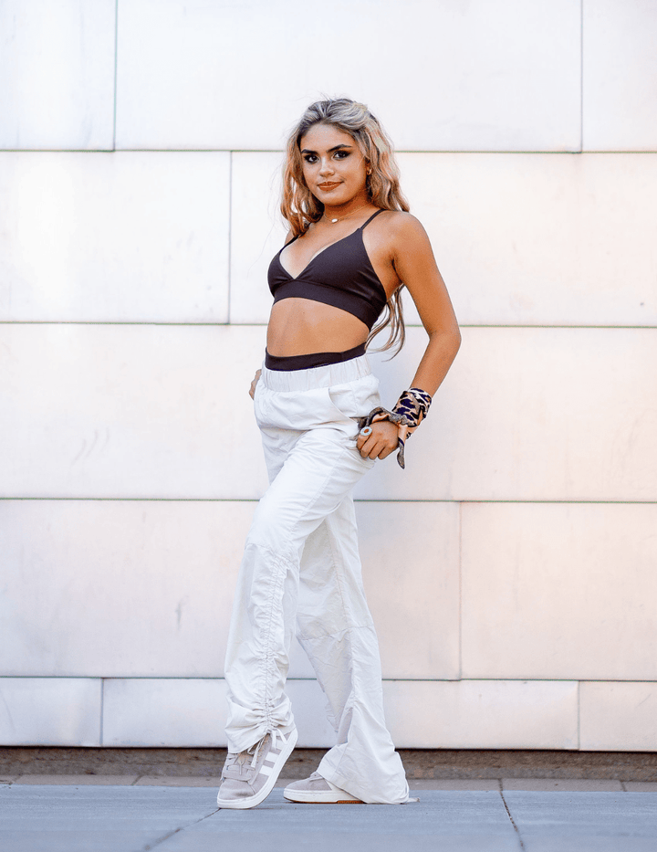 Woman wearing a black crop top and white pants standing against a light-colored wall.