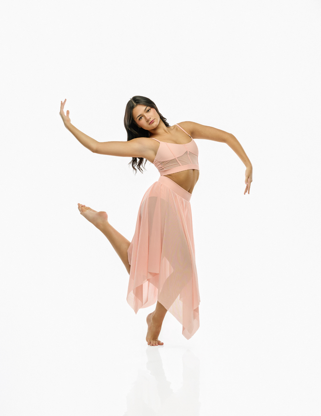 A woman in a light pink two-piece outfit consisting of a sheer crop top and a flowing high-low skirt, posing dynamically against a seamless white studio background.
