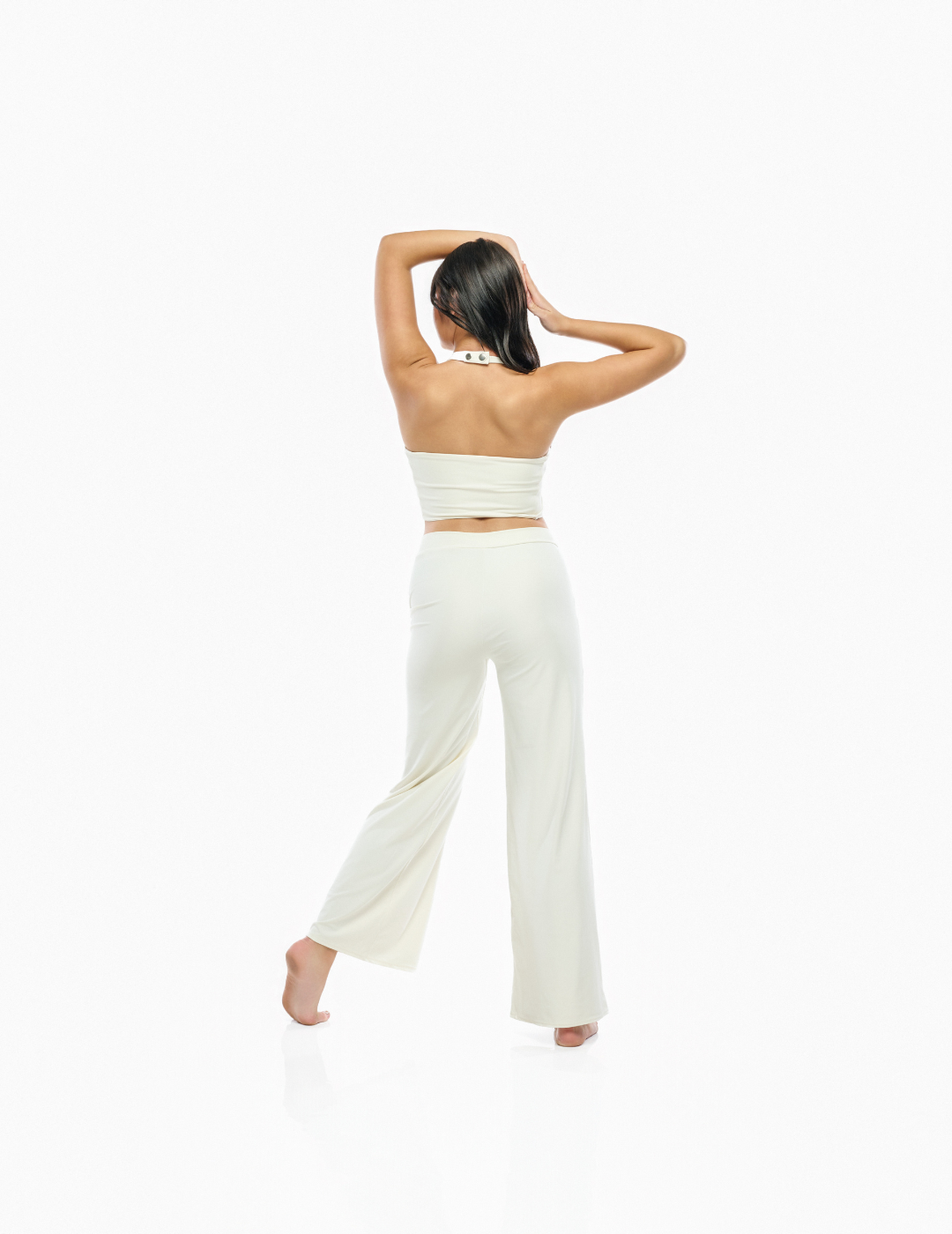 A woman is shown from behind wearing a matching two-piece loungewear set consisting of a strapless bandeau top and wide-leg pants, both in a warm off-white/cream color. She is standing barefoot against a seamless white studio background.
