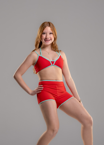 A young woman posing in a red two-piece swimsuit with light blue trim, consisting of a strappy bikini top and matching high-waisted shorts, against a solid gray background.