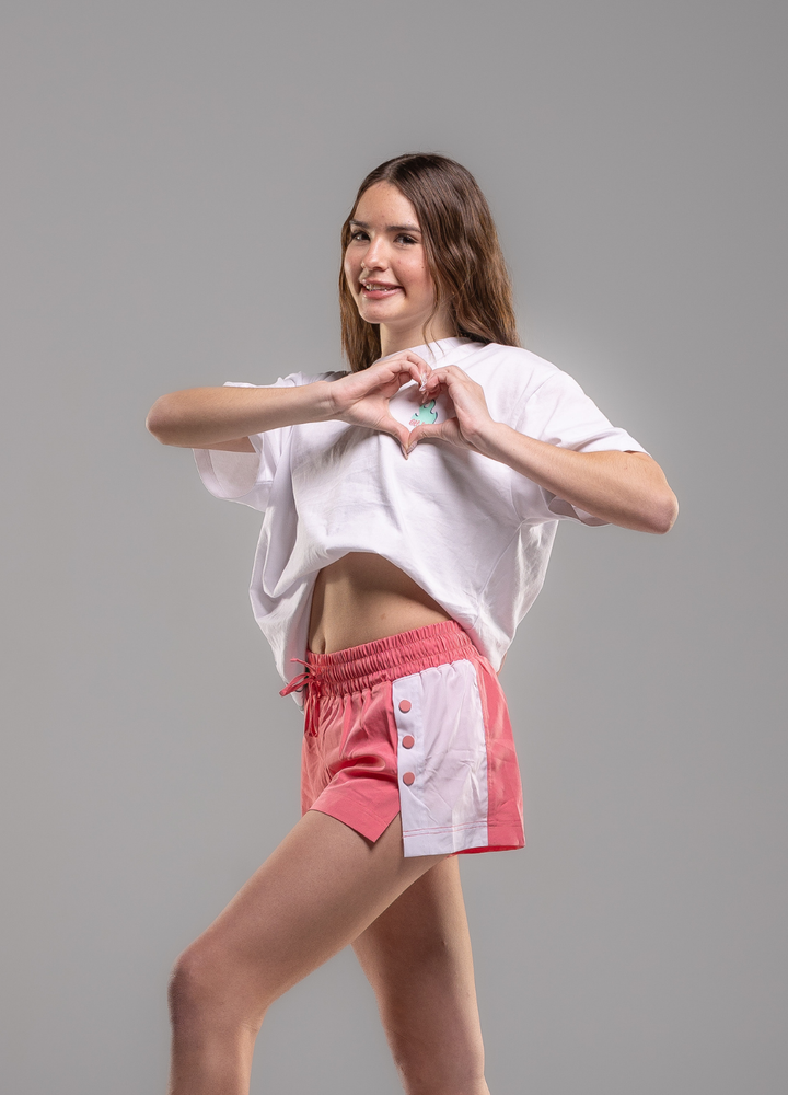 Woman wearing a white t-shirt and pink shorts making a heart shape with her hands on a gray background