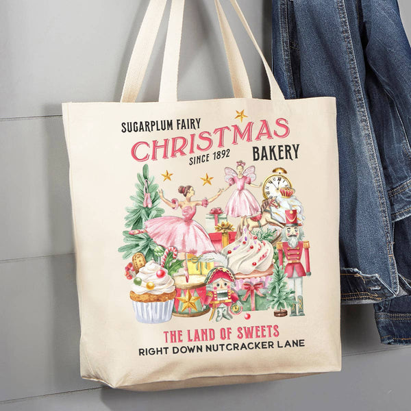 A cream canvas tote bag with a vintage-style Christmas bakery illustration, leaning against a gray paneled wall next to a blue denim jacket.