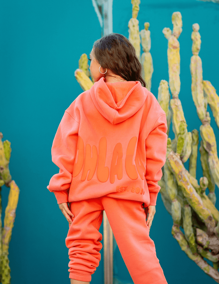 Person wearing a bright orange hoodie and pants with 'CHLAC' branding, standing in front of cacti.