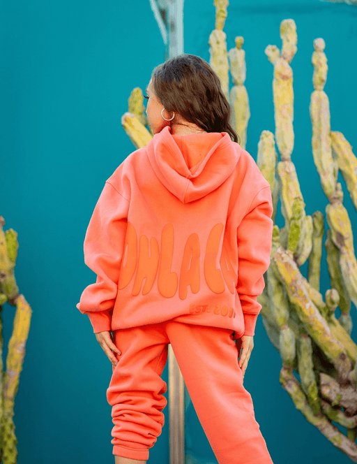 Person wearing a bright orange hoodie and pants with 'CHLAC' branding, standing in front of cacti.
