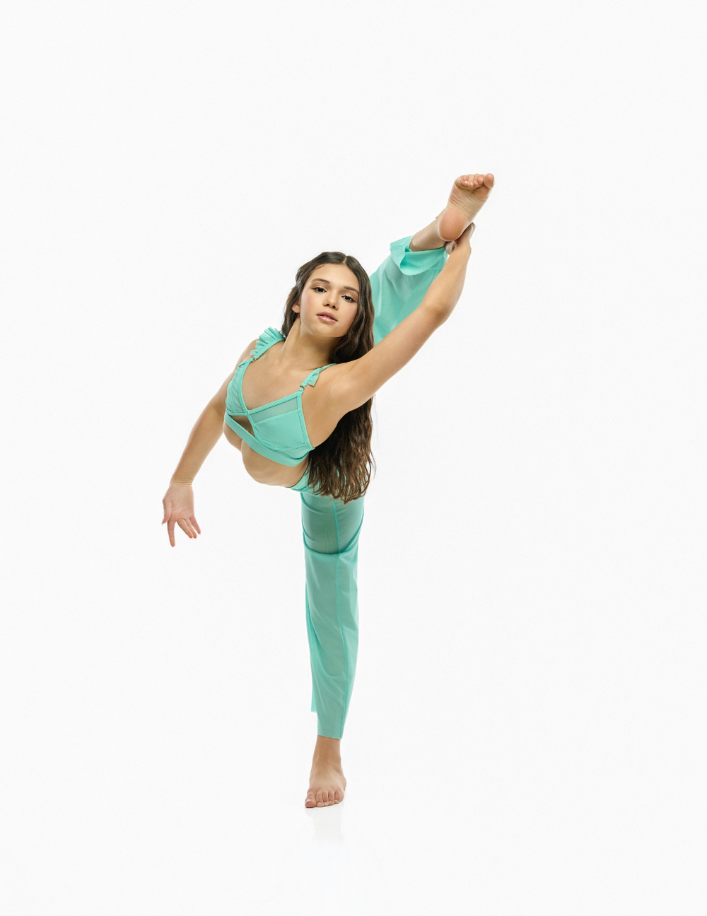 A woman in a flowing mint green dance dress poses in a high split leg stance against a plain white background.