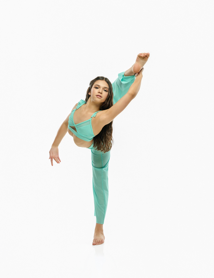 A woman in a flowing mint green dance dress poses in a high split leg stance against a plain white background.