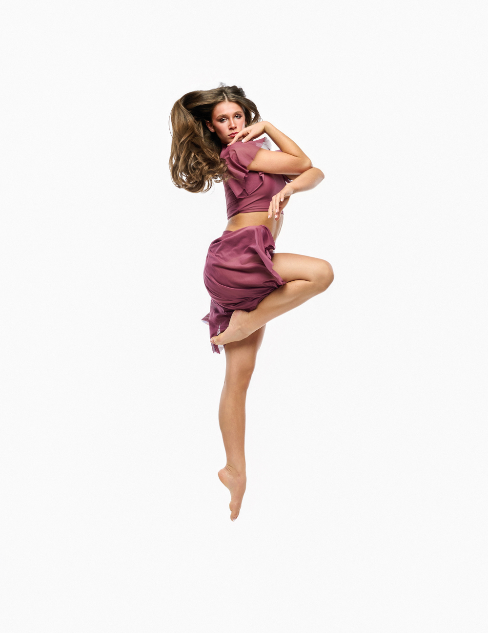 A woman in a matching mauve-purple two-piece outfit consisting of a short-sleeve crop top and a short skirt, captured mid-jump against a solid white background.