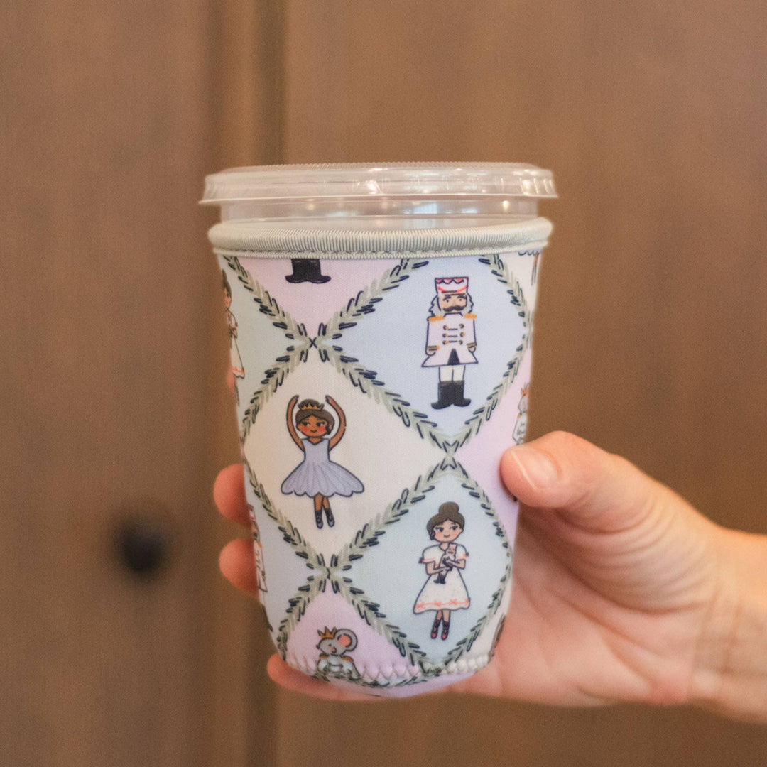 A hand holding a clear-lidded insulated coffee cup sleeve with a white background and a repeating pattern of illustrated ballerina and nutcracker figures inside a diamond lattice design, set against a warm brown wooden door background.