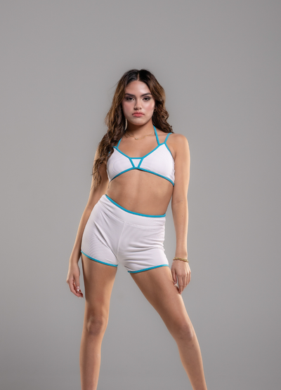 A woman modeling a white two-piece athletic set with bright turquoise blue trim, consisting of a strappy sports bra and matching high-waisted shorts, against a solid gray studio background.