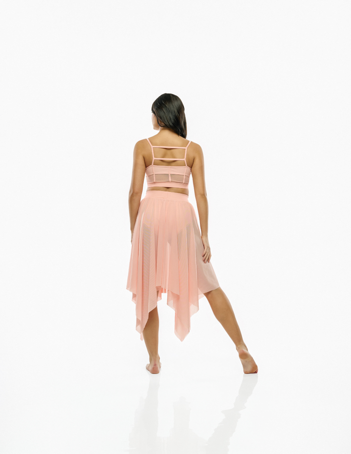 A woman viewed from behind wearing a light peach-pink two-piece set consisting of a strappy crop top and a matching high-low tulle skirt, set against a seamless white studio background.