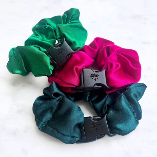 Three satin scrunchies in green, pink, and teal with black plastic clips on a white background.